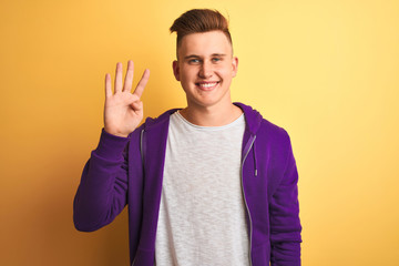 Young handsome man wearing purple sweatshirt standing over isolated yellow background showing and pointing up with fingers number four while smiling confident and happy.