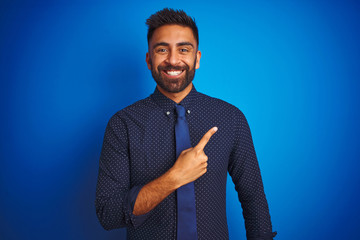 Wall Mural - Young indian businessman wearing elegant shirt and tie standing over isolated blue background cheerful with a smile of face pointing with hand and finger up to the side with happy and natural