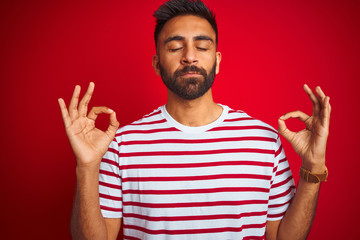 Wall Mural - Young indian man wearing striped t-shirt standing over isolated red background relax and smiling with eyes closed doing meditation gesture with fingers. Yoga concept.