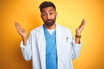 Wall Mural - Young indian doctor man standing over isolated yellow background clueless and confused expression with arms and hands raised. Doubt concept.