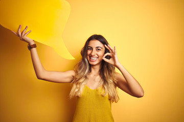 Wall Mural - Young beautiful woman holding speech bubble over yellow isolated background doing ok sign with fingers, excellent symbol