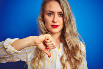 Wall Mural - Young beautiful woman wearing striped shirt standing over blue isolated background with angry face, negative sign showing dislike with thumbs down, rejection concept