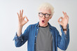 © Krakenimages.com - Young albino blond man wearing denim shirt and glasses over isolated white background looking surprised and shocked doing ok approval symbol with fingers. Crazy expression