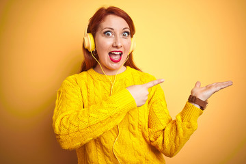 Sticker - Young redhead woman listening to music using headphones over yellow isolated background amazed and smiling to the camera while presenting with hand and pointing with finger.