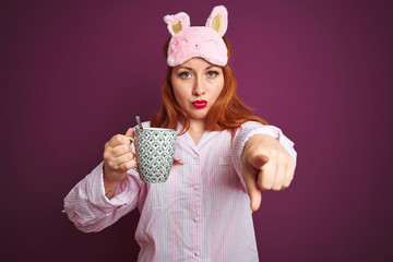 Sticker - Young redhead woman wearing pajama drinking coffee over purple isolated background pointing with finger to the camera and to you, hand sign, positive and confident gesture from the front