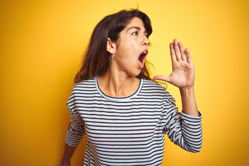 Wall Mural - Young beautiful woman wearing stripes t-shirt standing over yelllow isolated background shouting and screaming loud to side with hand on mouth. Communication concept.