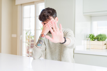 Poster - Young man wearing casual sweater sitting on white table covering eyes with hands and doing stop gesture with sad and fear expression. Embarrassed and negative concept.