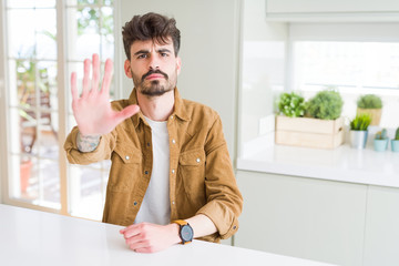 Sticker - Young man wearing casual jacket sitting on white table doing stop sing with palm of the hand. Warning expression with negative and serious gesture on the face.