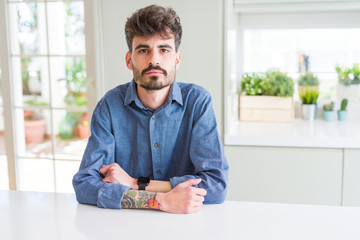 Poster - Young man wearing casual shirt sitting on white table Relaxed with serious expression on face. Simple and natural looking at the camera.