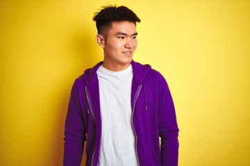 Young asian chinese man wearing purple sweatshirt standing over isolated yellow background looking away to side with smile on face, natural expression. Laughing confident.
