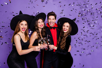 Canvas Print - Photo of three witch ladies and wizard guy at helloween event drink golden wine glitter in air wear black dresses caps suit and long coat isolated purple color background