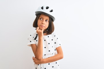 Poster - Beautiful child girl wearing security bike helmet standing over isolated white background with hand on chin thinking about question, pensive expression. Smiling with thoughtful face. Doubt concept.