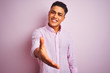 Young brazilian man wearing shirt standing over isolated pink background smiling friendly offering handshake as greeting and welcoming. Successful business.