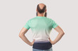 backside portrait of middle aged bald man with beard in light green t-shirt standing with hands on waists. indoor studio shot, isolated on grey background.