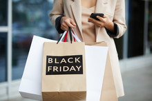 Mid-section portrait of stylish young woman holding shopping bags with Black Friday and texting on the go while leaving mall in sale season, copy space
