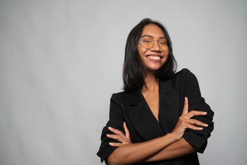 Portrait of attractive young Asian smiling woman in elegant black blazer and glasses posing over light gray wall background. Mock up copy space