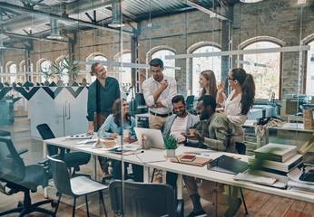 Group of young modern people in smart casual wear communicating and using modern technologies while working in the office