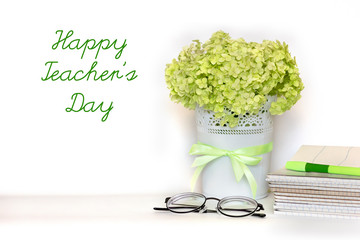 Happy Teacher's day greeting card - bouquet of light green hydrangea branch in flower pot with satin ribbon, pile of notebooks, glasses and pen on white background. International school Teacher Day.