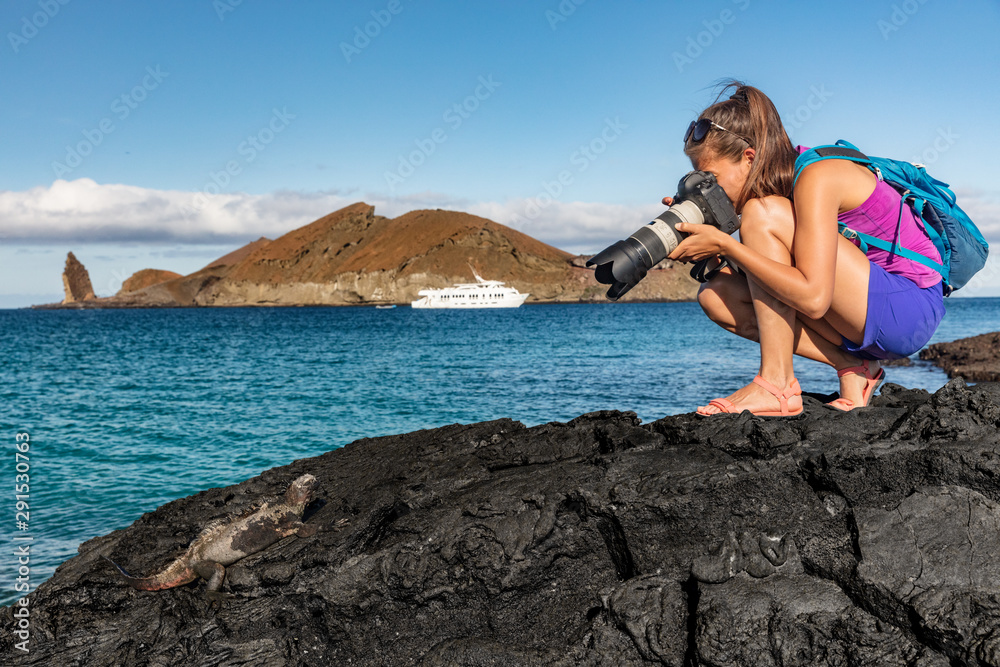 Galapagos tourist photographing marine iguana on Santiago Island in Galapagos Islands. Cruise ship and Pinnacle Rock and Bartolome Island in background. Famous Galapagos cruise ship tour destination. - obrazy, fototapety, plakaty 