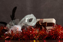 A Gray Gift Box With A Brown Bow Lies In Purple Tinsel With Carnival Mask