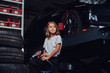 Little cute girl is posing for photographer in dark auto workshop with pneumatic drill in hands.