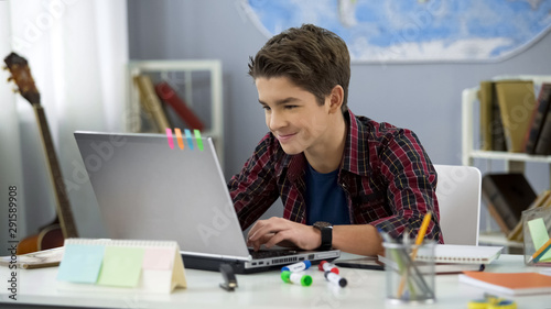 Fotografía Smiling teen male chatting with friends in social networks sitting front laptop