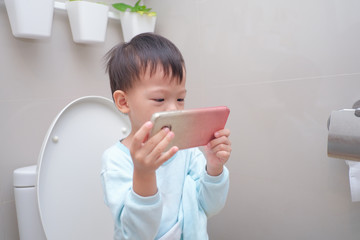 Cute little Asian 3 - 4 years old toddler boy kid using mobile phone while sitting on toilet seat in bathroom at home, Screentime & Potty Training & young children concept, photo in real life interior