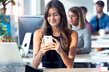 Smiling young businesswoman using her smartphone while working with computer in the modern startup office.