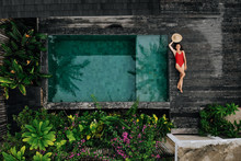 Aerial Drone Photo Of Happy Woman In Red Swimsuit Relaxing Near Private Pool With Flowers And Greenery Around, Bali. Tropical Background And Travel Concept.