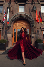 Young Beautiful Stunning Woman In Red Luxury Evening Dress With Flying Hem Walking City Street On A Sunny Evening. Elegant Lady With Makeup And Wavy Brunette Hair. Full Length Fashion Portrait