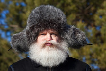 Funny Senior Caucasian Man In Grey Fur Hat Making Grimaces. Closeup Sunny Portrait Of Elderly Stylish Person In Black Flight (bomber) Jacket At Nature Against Dark Forest Background.