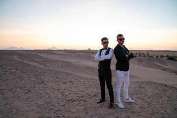 Two handsome friends in sunglasses and elegant closes, one in white shirt other in black standing with crossed hands among the desert, sunset on the background