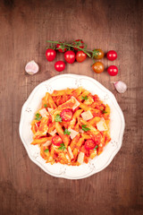 Wall Mural - Penne pasta with chicken, tomato sauce and parsley, shot from above on a dark rustic wooden background with ingredients