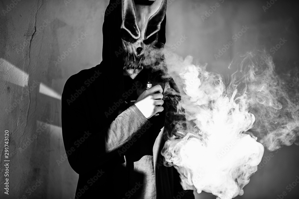 Vape man in devil mask and a hood smoking an electronic cigarette outdoors.  Puffs of steam. Bad habit that is harmful to health. Vaping activity. Black  and white. Stock Photo | Adobe