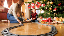 Cheerful Smiling Boy With Young Mother Building Railroad Around Christmas Tree At Living Room. Child Receiving Presents And Toys On New Year Or Xmas