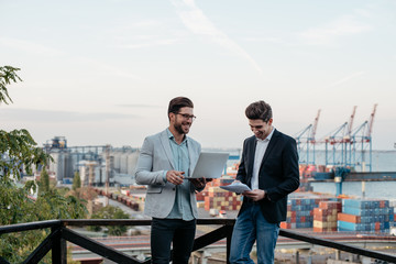 Wall Mural - Two business partners smiling and discussing the meeting with laptop and contract, outdoor on background of the seaport