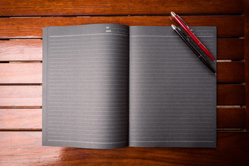 A large black writing pad on which are two office pens in black and red located on a wooden table