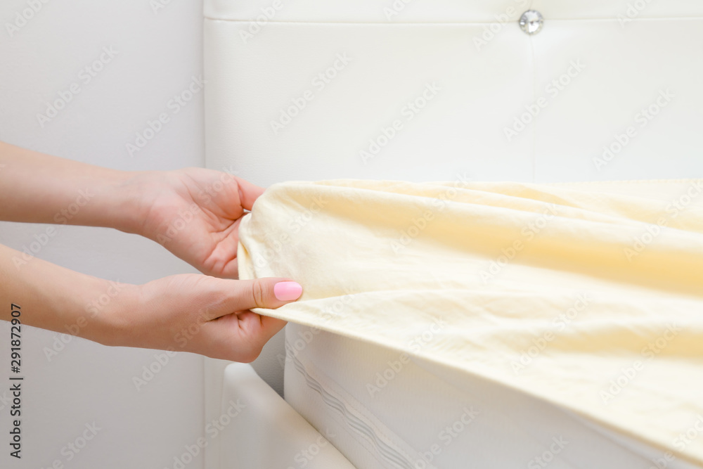 Woman's hands changing light yellow sheet on white mattress. Regular bed linen change. Closeup. Side view. - obrazy, fototapety, plakaty 