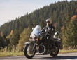 Handsome bearded biker in black leather jacket and sunglasses sitting on cruiser motorcycle on country roadside, on blurred background of foggy green hills covered with dense spruce forest.