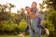 Portrait of cheerful spouses with brunet hair piggyback wearing denim jeans in park outside