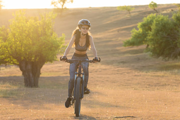 Girl on a mountain bike on offroad, beautiful portrait of a cyclist at sunset, Fitness girl rides a modern carbon fiber mountain bike in sportswear, a helmet, glasses and gloves.	