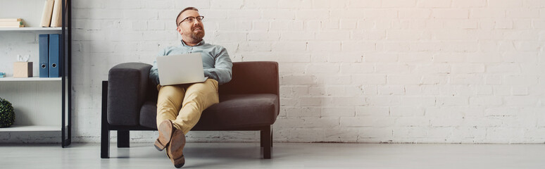 panoramic shot of handsome businessman in shirt holding laptop and looking away