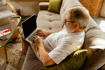 Senior man working with laptop at home - concept of home studying. Caucasian male model sitting on sofa and doing his homework while serfing in internet, watching cinema or webinar, online lessons.