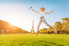 Happy Asian Woman Traveler Jumping With French Flag At Mars Field At The Background Of Majestic Eiffel Tower. Tourism And Lifestyle In France And Paris. Vacation In Europe.