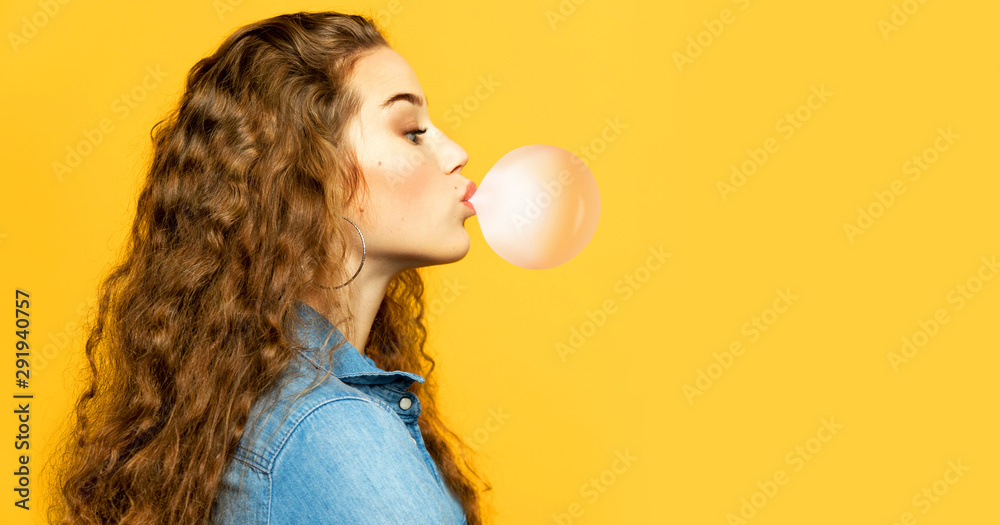 Portrait of funny young model chewing bubblegum. Attractive woman posing in profile in studio on yellow background. Copy space in right side. Emotions and lifestyle concept - obrazy, fototapety, plakaty 