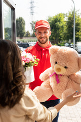  selective focus of cheerful delivery man in cap holding teddy bear and flowers near woman