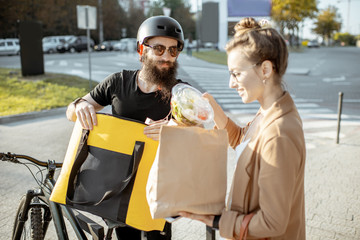 Male courier delivering food on a bicycle, giving bag with fresh takeaway lunch to a young business woman outdoors. Restaurant food delivery concept