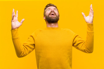 Wall Mural - young blonde man feeling happy, amazed, lucky and surprised, celebrating victory with both hands up in the air against orange wall