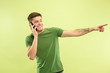 © master1305 - Caucasian young man's half-length portrait on green studio background. Beautiful male model in shirt. Concept of human emotions, facial expression, sales, ad. Talking on phone and pointing, smiling.