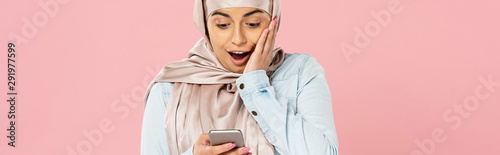 Fotomural shocked muslim girl in hijab using smartphone isolated on pink